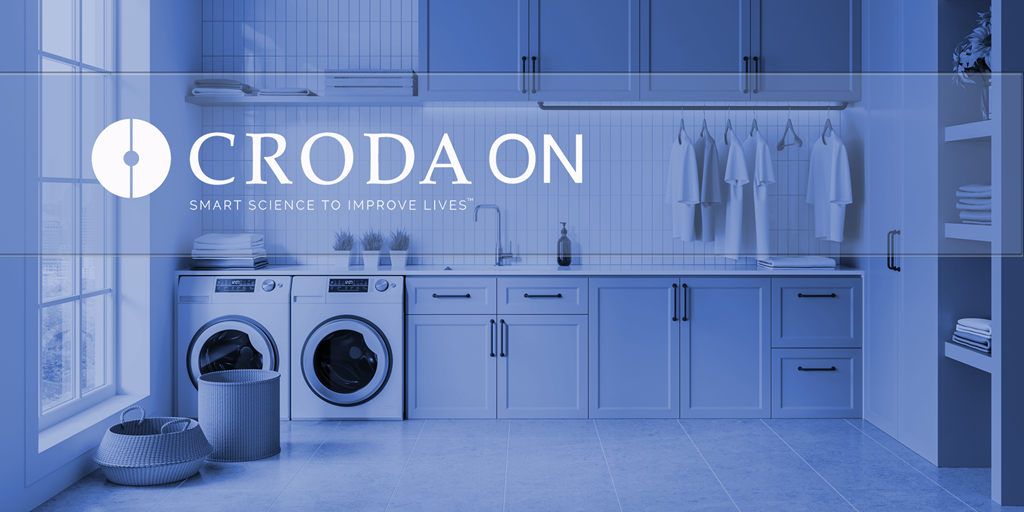 A very clean laundry room with washing machine and tumble dryer, laundry basket, clean clothes hanging up and a sink. The words "Croda On - Smat science to improve lives" are written across it.